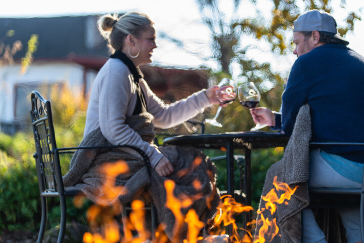 A cold winter day shows a couple toasting glasses of wine at a winery's outdoor seating section in front of a fire pit. The man is wearing a navy blue pullover sweater, jeans, and a baseball cap. The woman is wearing a white cashmere sweater, dark scarf, and hoop earrings. Both have a blanket across their laps for extra warmth. 