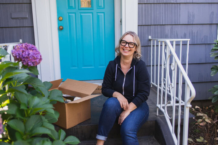 A blonde woman sits on the front stoop of her doorstep in front of a bright blue door. She is wearing a black zip up hoodie and dark jeans. She is smiling and sitting next to wine bottles delivered in eco-friendly packaging.