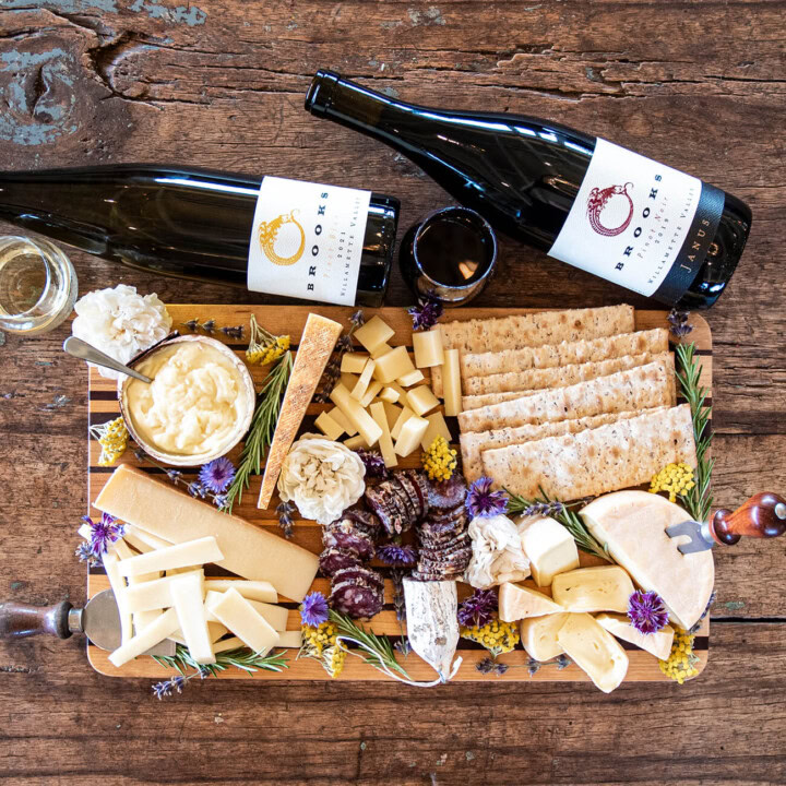 A rustic wooden table lifts a bountiful board filled with various cheeses, charcuterie, crackers, and decorative flowers alongside two bottles of wine laying length-wise along the top of the board.