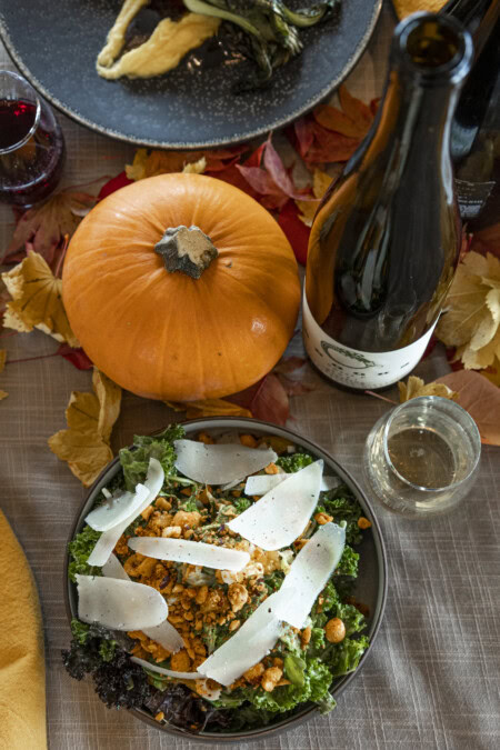 A gray linen tablecloth is decorated for Thanksgiving with red and yellow leaves and orange pumpkins alongside a bottle of Riesling and a bountiful bowl of dark greens, crispy bread crumbs, and thick shavings of parmesan.