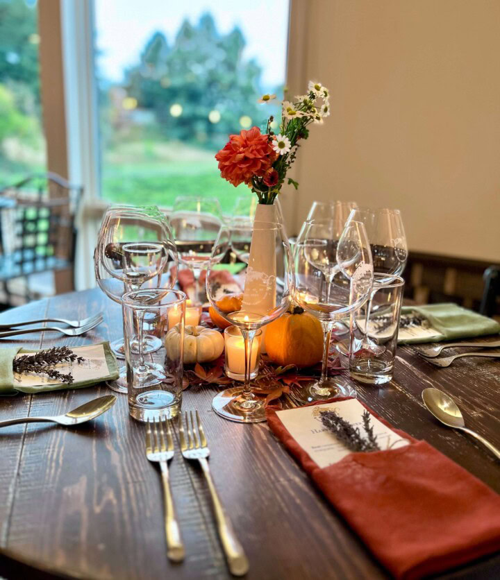 A wooden table is set for entertaining guests at Friendsgiving with dark orange cloth napkins folded to hold a menu and sprig of lavender with the center of the table displaying votive candles, dried leaves, small pumpkins, and a thin white vase holding orange flowers. 