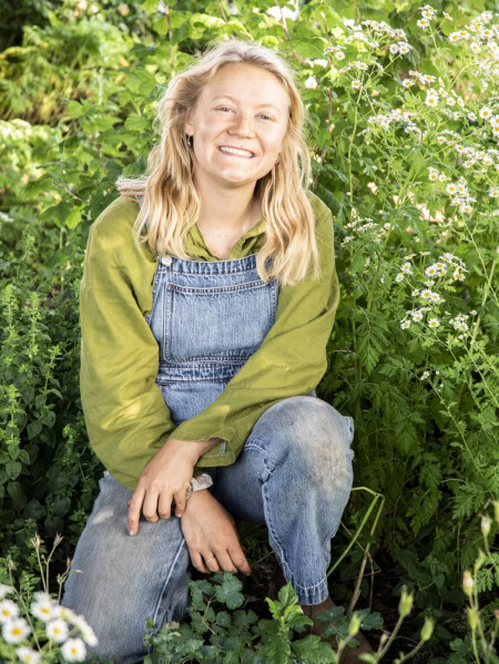 Shannon Mayhew, the Biodynamic Estate Gardener at Brooks Winery in Oregon's Willamette Valley, is shown kneeling in front of lush greenery and white flowers while wearing an avocado green linen shirt and denim overalls.