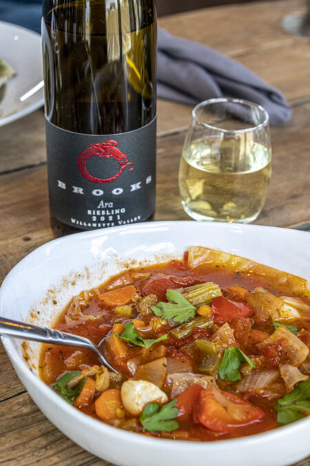 A wooden table is set with a white bowl of beef and winter vegetable strew paired with a crisp dry Riesling in a stemless glass next to the wine bottle with a black label. 