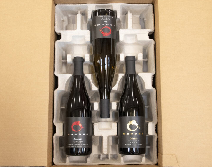 Three bottles of wine are laying horizontally within a cardboard shipping cradle and box. 