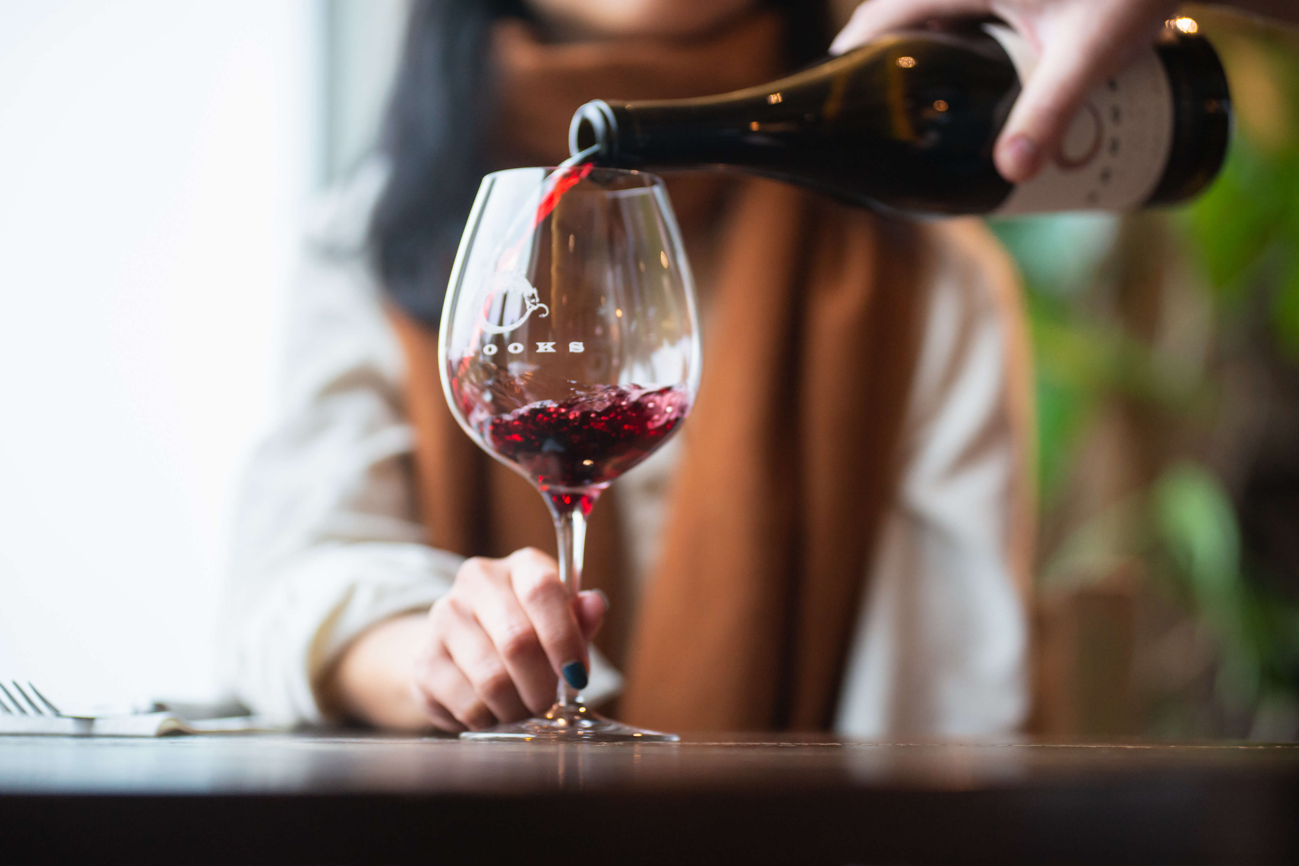A dark-haired woman wearing a white blouse and brown scarf is poured a glass of Pinot Noir as she holds the glass by the stem in observance of wine etiquette.